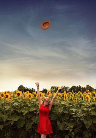 Woman in red dress in a field of sunflowers throwing her hat into the air.